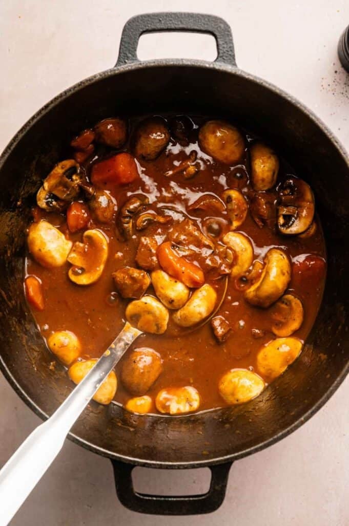 A black pot filled with hearty Beef and Guinness Stew, featuring a rich brown sauce, whole mushrooms, chunks of carrots, and a metal spoon resting inside.