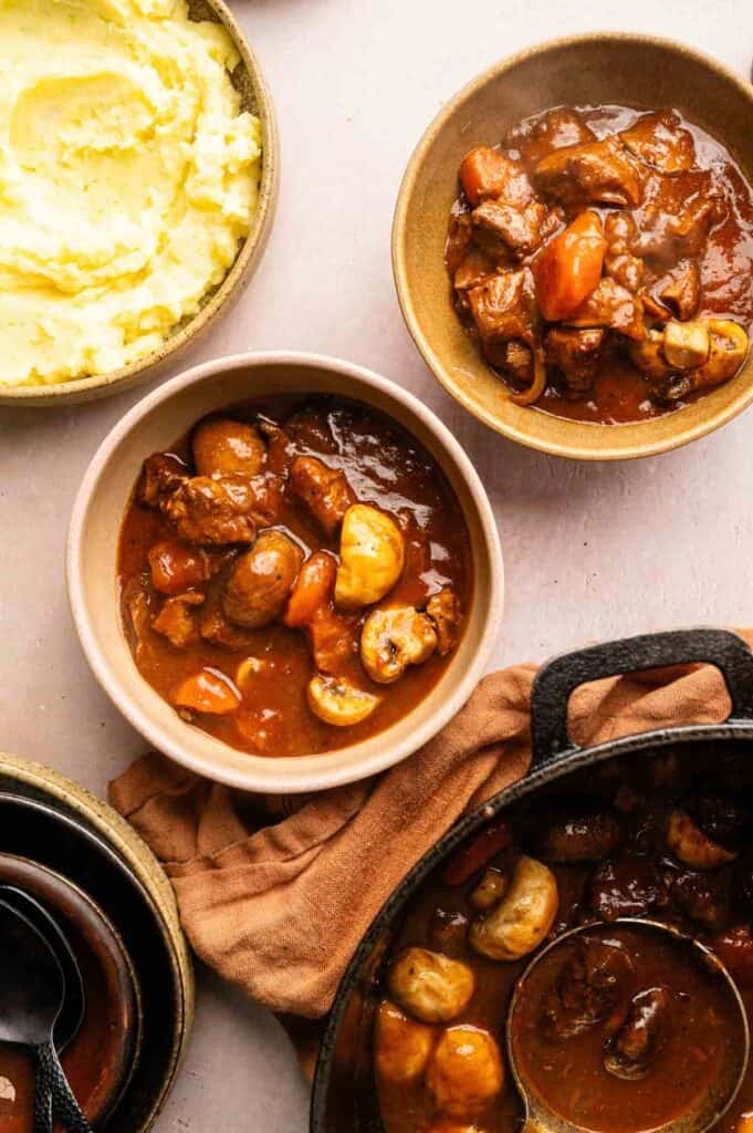 Bowls of Beef and Guinness Stew with carrots and mushrooms are arranged next to a bowl of mashed potatoes and a pot of stew with a ladle.