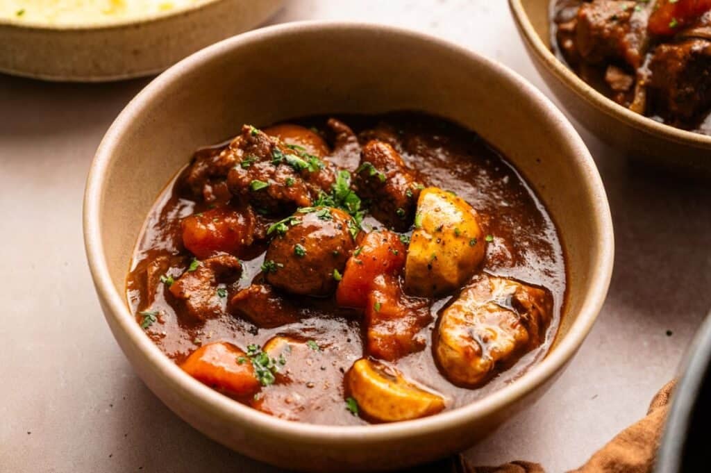 A bowl of Beef and Guinness Stew with chunks of beef, carrots, potatoes, and mushrooms in a thick brown broth, garnished with chopped herbs.