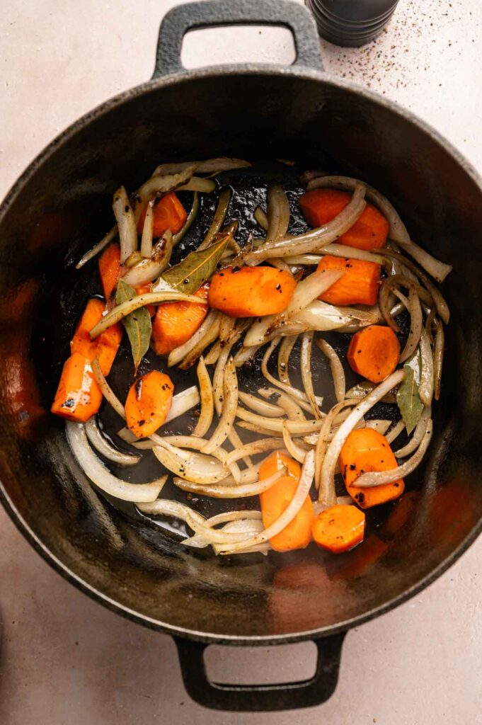 Sliced onions, carrot chunks, and bay leaves sautéing in a black cast iron pot—the perfect aromatic base for a hearty Beef and Guinness Stew.