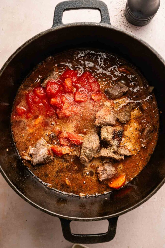 A black pot filled with simmering Beef and Guinness Stew, chunks of meat, diced tomatoes, and visible seasonings on a light surface.