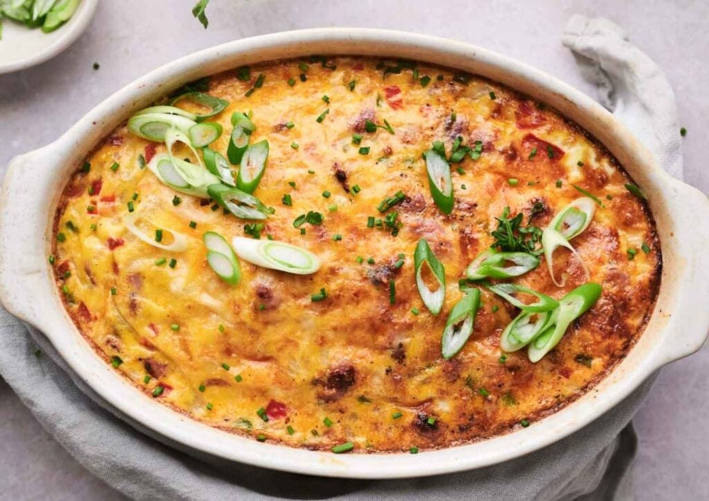 Baked breakfast casserole topped with sliced green onions and herbs, in a white oval dish on a gray cloth, viewed from above.