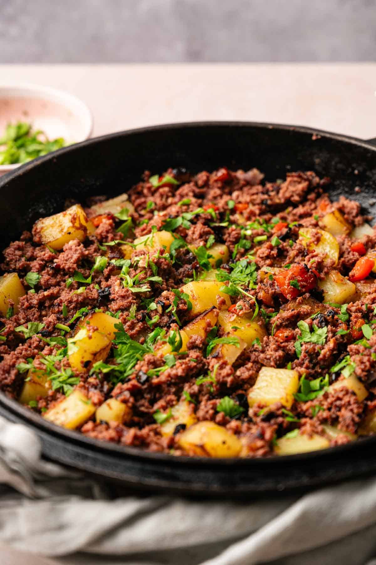 A skillet filled with cooked ground beef, diced potatoes, tomatoes, and garnished with chopped herbs&mdash;a savory twist on classic Corned Beef Hash.