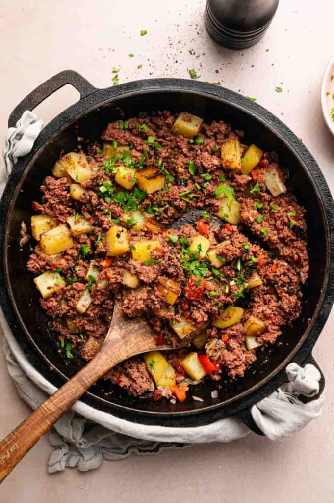 A cast iron skillet filled with corned beef hash&mdash;ground beef, diced potatoes, and chopped vegetables&mdash;garnished with fresh herbs, with a wooden spoon resting in the pan.
