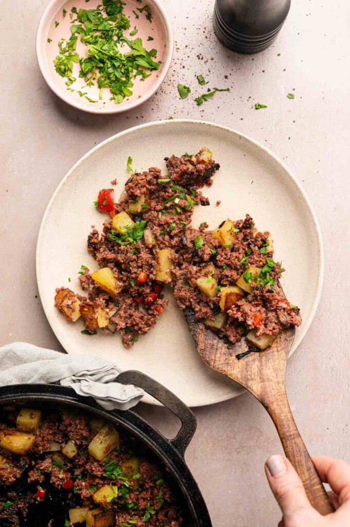 A person serves corned beef hash with diced potatoes and peppers from a skillet onto a plate, with chopped herbs in a small bowl nearby.