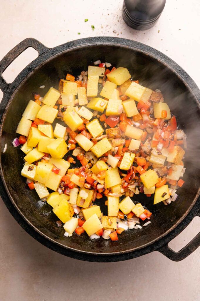 Diced potatoes, carrots, onions, and red bell peppers are sautéing in a black cast iron skillet, creating the perfect base for a classic Corned Beef Hash.