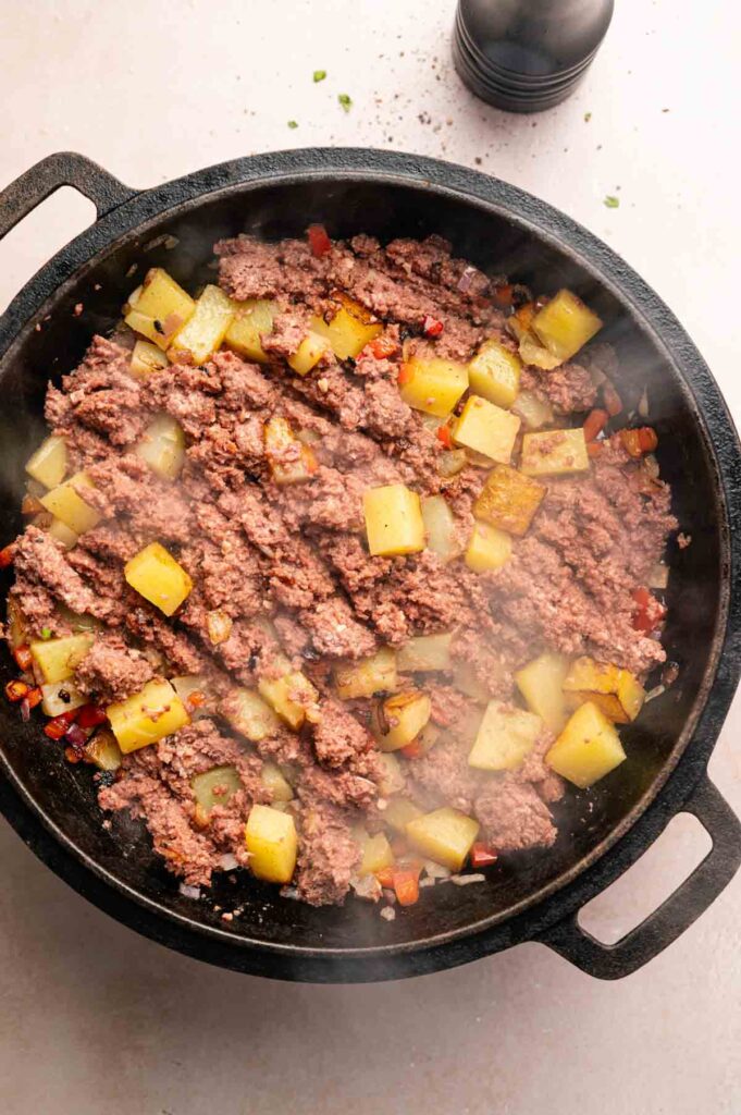 A cast iron skillet filled with cooked ground beef, diced potatoes, and small pieces of bell pepper seen from above, reminiscent of a classic Corned Beef Hash.
