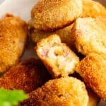 A close-up of several golden-brown Ham Croquettes on a plate, with one croquette broken in half to reveal a creamy filling studded with bits of ham.