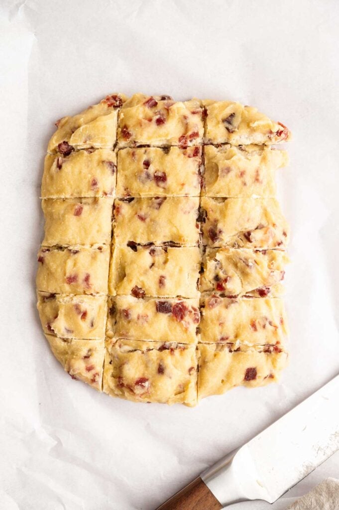 Square dough with bacon bits is cut into 16 pieces on parchment paper, with a knife placed next to it—reminiscent of preparing delicious ham croquettes for baking or frying.