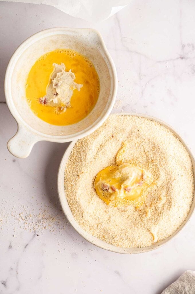 A bowl with beaten eggs and a piece of food beside a bowl of breadcrumbs, with a Ham Croquettes-coated item being breaded on a white surface.