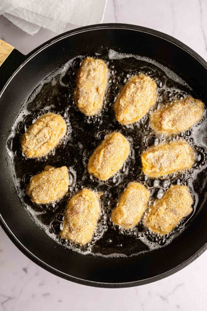 Ham croquettes are frying in hot oil in a black skillet on a marble surface. The breaded croquettes sizzle to golden perfection, promising a savory treat.