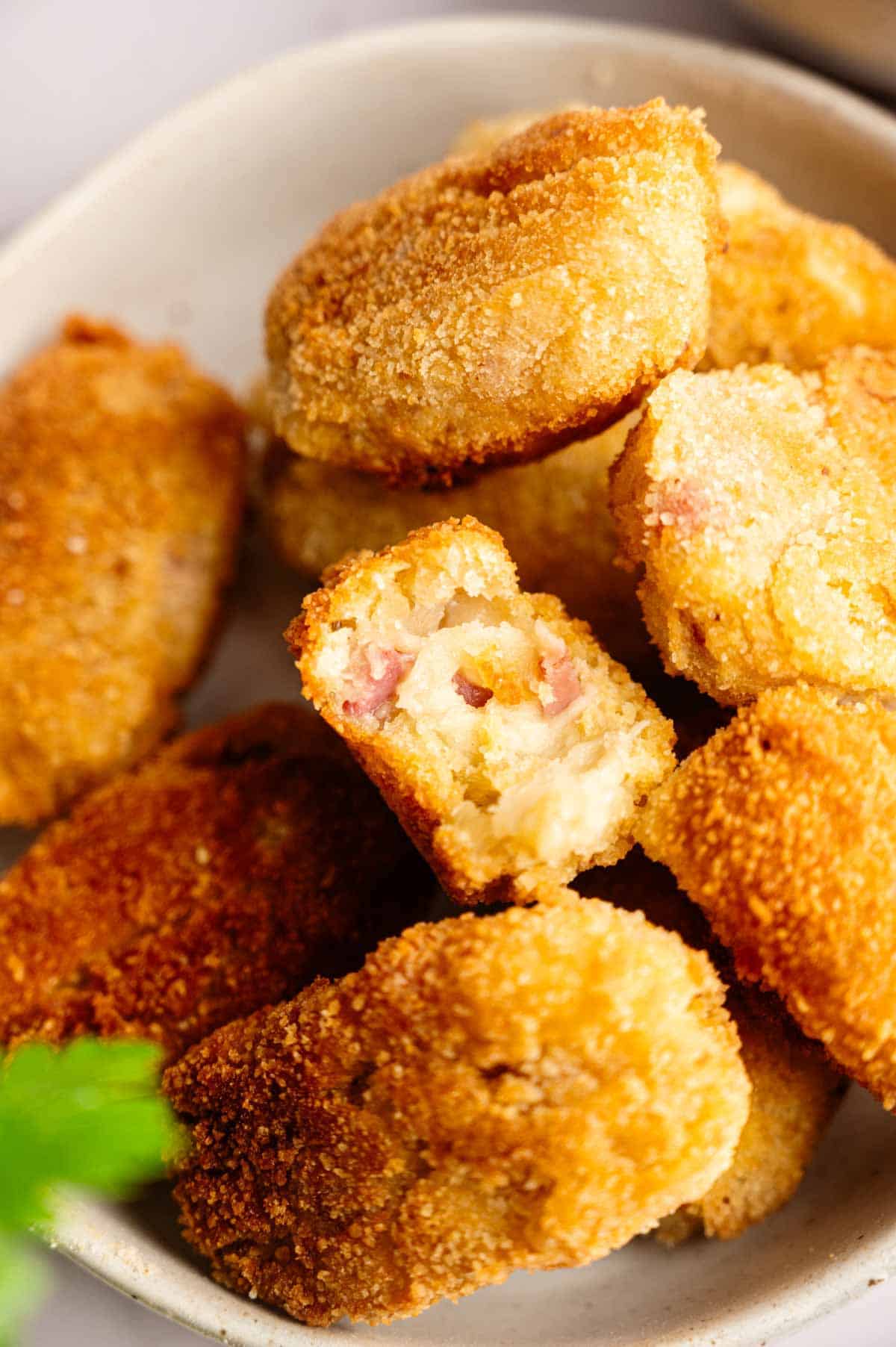 A bowl of golden-brown Ham Croquettes, with one croquette broken in half to reveal a creamy filling speckled with bits of ham inside.