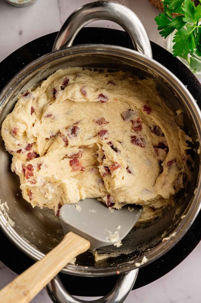 A bowl of thick, mixed batter for Ham Croquettes, with pieces of ham or bacon visible, being stirred with a spatula.