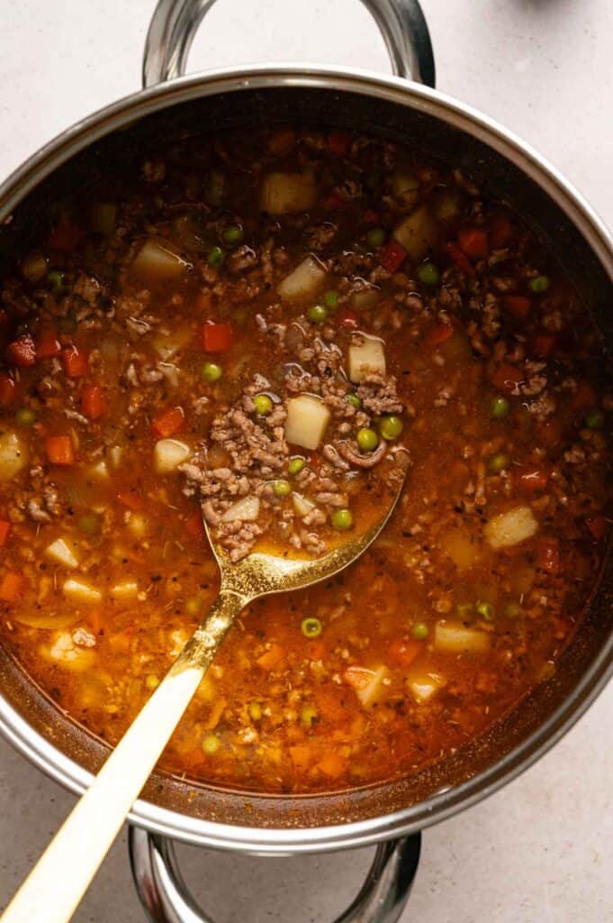A pot of Hamburger Soup with ground beef, potatoes, peas, and carrots, with a ladle resting inside.