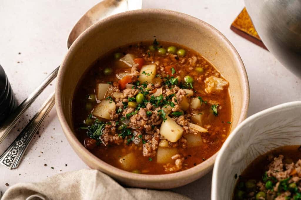 A bowl of hearty hamburger soup with ground meat, potatoes, peas, carrots, and broth, garnished with chopped herbs, rests on a light tabletop with a spoon nearby.