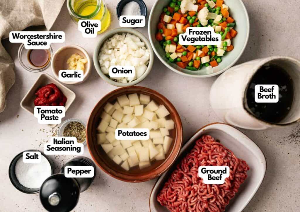 Top-down view of labeled Hamburger Soup ingredients, including ground beef, potatoes, onion, garlic, frozen vegetables, beef broth, tomato paste, Worcestershire sauce, seasonings, salt, pepper, sugar, and olive oil.