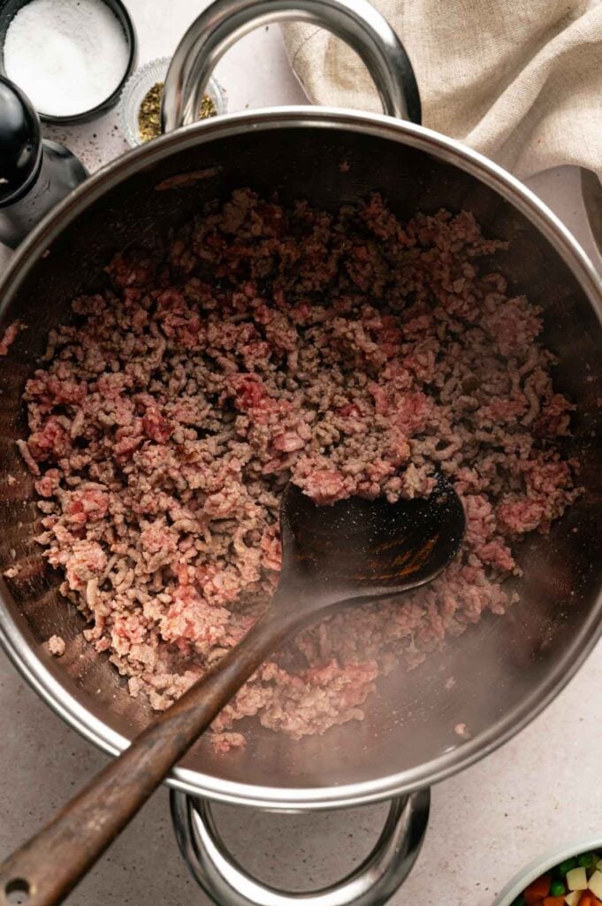 Ground beef is being browned in a large metal pot, stirred with a wooden spoon—the perfect savory base for comforting Hamburger Soup.