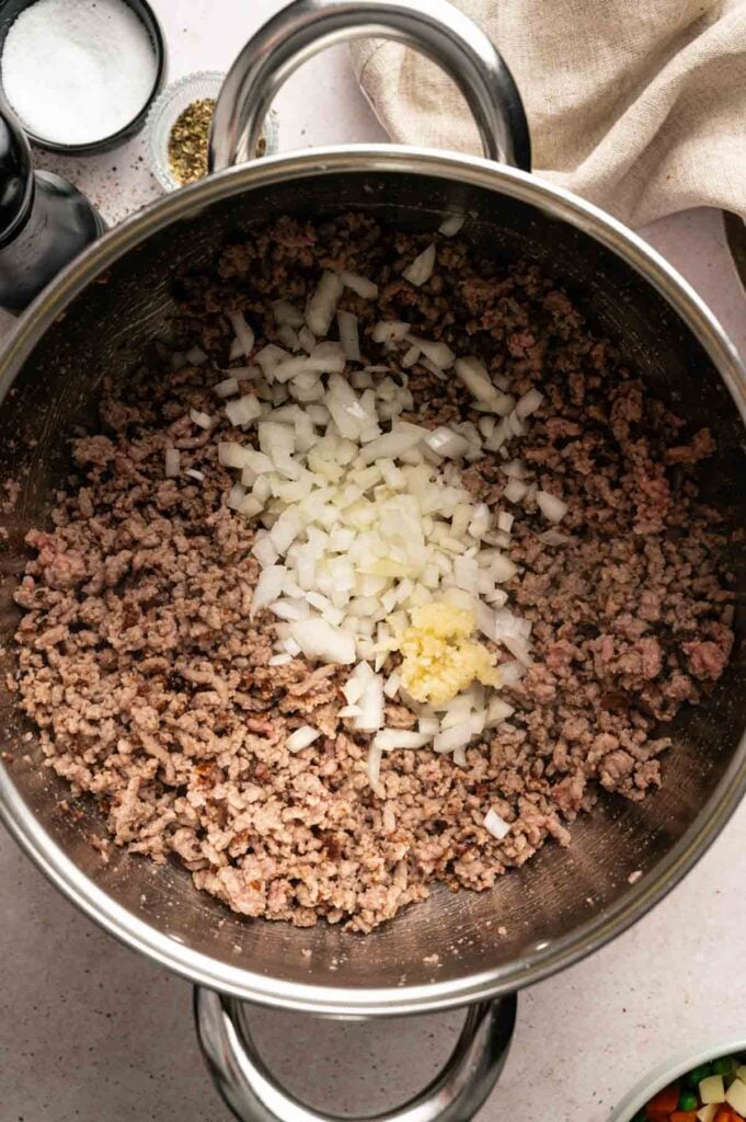 Chopped onions and minced garlic are added to browned ground beef in a large stainless steel pot, forming the hearty base for a flavorful Hamburger Soup, ready to be mixed and cooked further.
