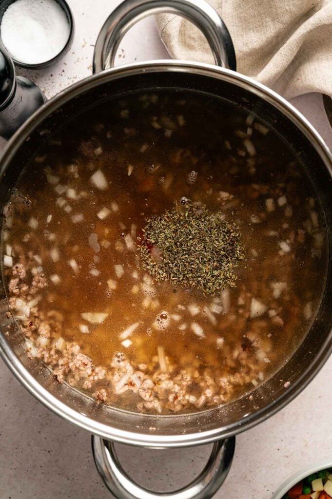 A stainless steel pot contains broth with ground meat, chopped onions, and a pile of dried herbs in the center, ready to be mixed—perfect for making hearty Hamburger Soup.
