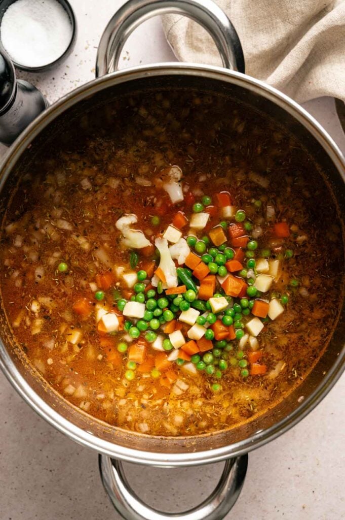 A pot of hamburger soup with peas, carrots, potatoes, onions, and broth, partially cooked and still simmering on a stove.