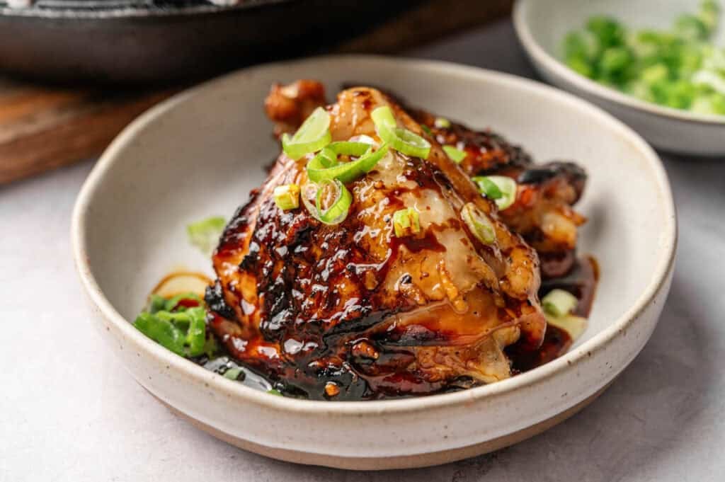 A bowl with glazed Honey Garlic Chicken Thighs topped with sliced green onions, served with extra green onions on the side.