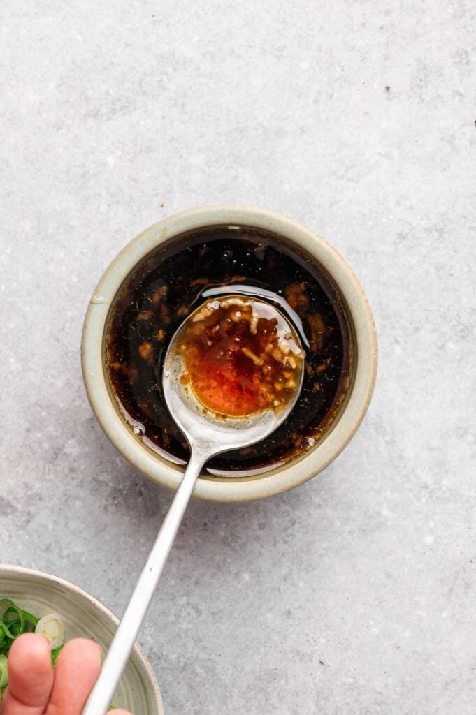 A metal spoon rests in a small bowl filled with a dark, oily sauce containing visible bits of garlic or seasoning—perfect for drizzling over Honey Garlic Chicken Thighs.