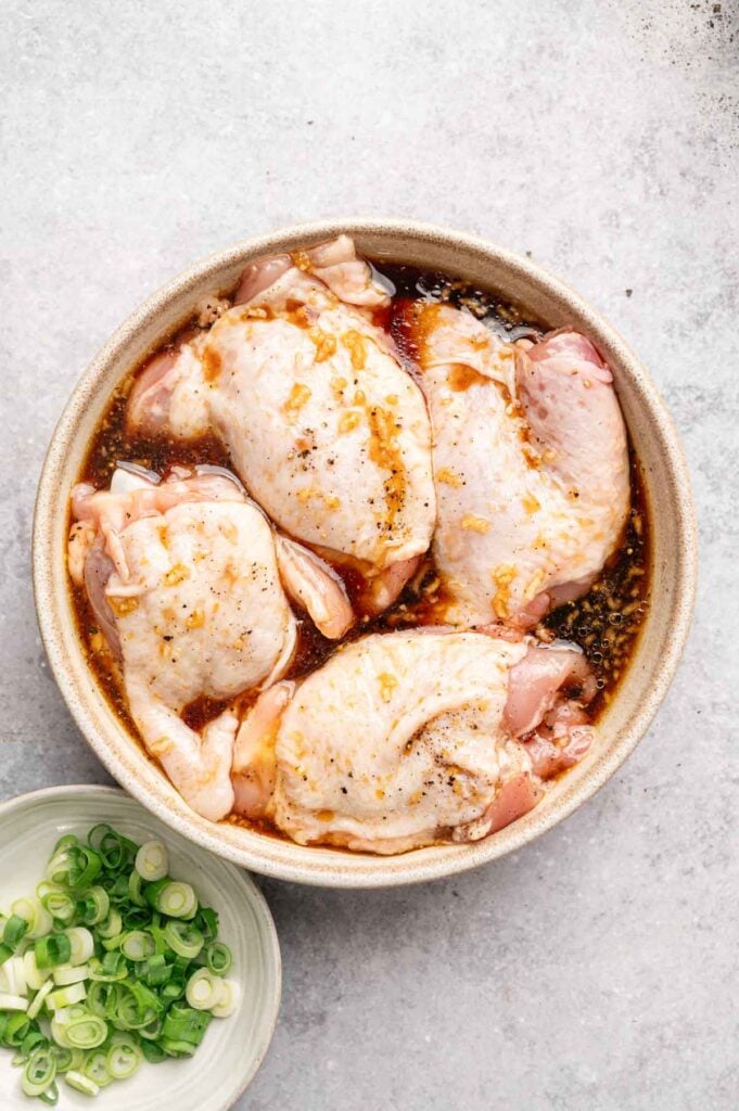Four raw chicken thighs marinating in a bowl of soy-based sauce, perfect for Honey Garlic Chicken Thighs, with a small bowl of chopped green onions beside them on a light surface.