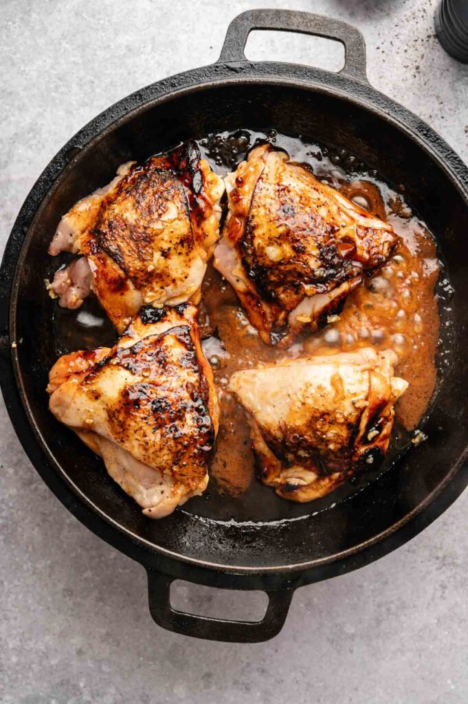 Four browned Honey Garlic Chicken Thighs in a cast iron skillet with pan juices, placed on a light gray surface.