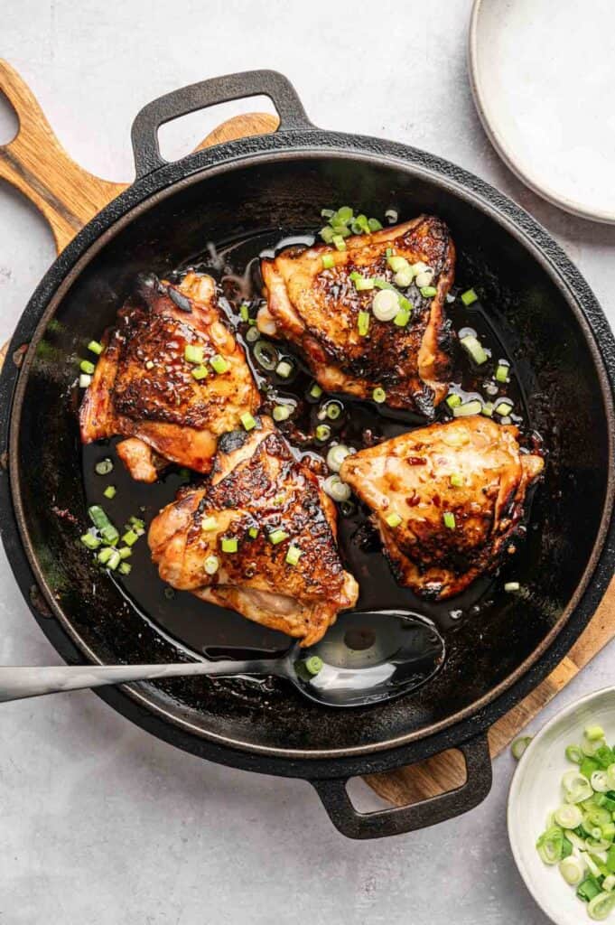 Four browned Honey Garlic Chicken Thighs with sauce and chopped green onions in a black cast iron skillet, with a spoon and a wooden handle visible.