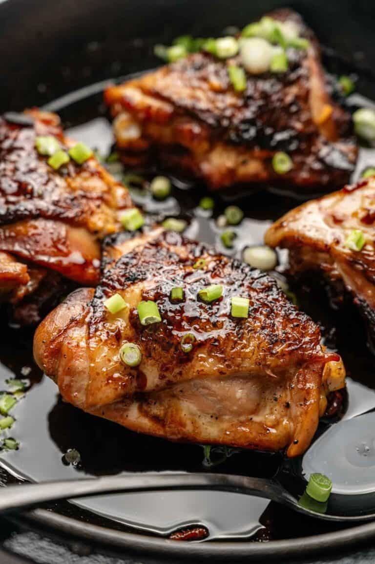 Close-up of Honey Garlic Chicken Thighs seared in a skillet, topped with chopped green onions and sitting in a dark, glossy sauce.