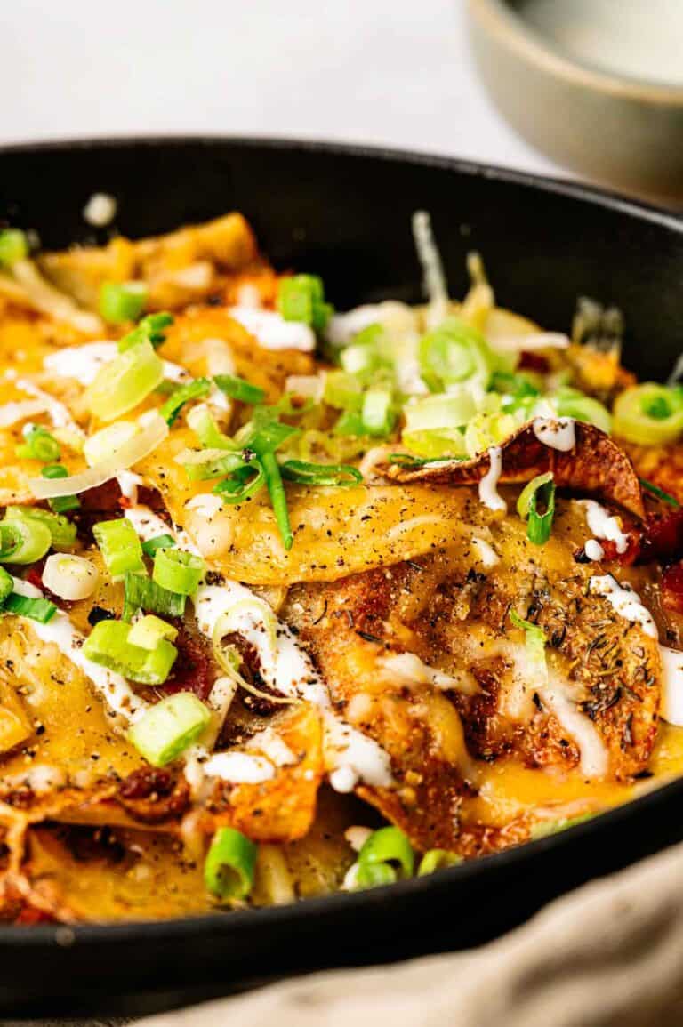 A close-up of a skillet filled with Irish Nachos topped with melted cheese, sliced green onions, black pepper, and a drizzle of white sauce.