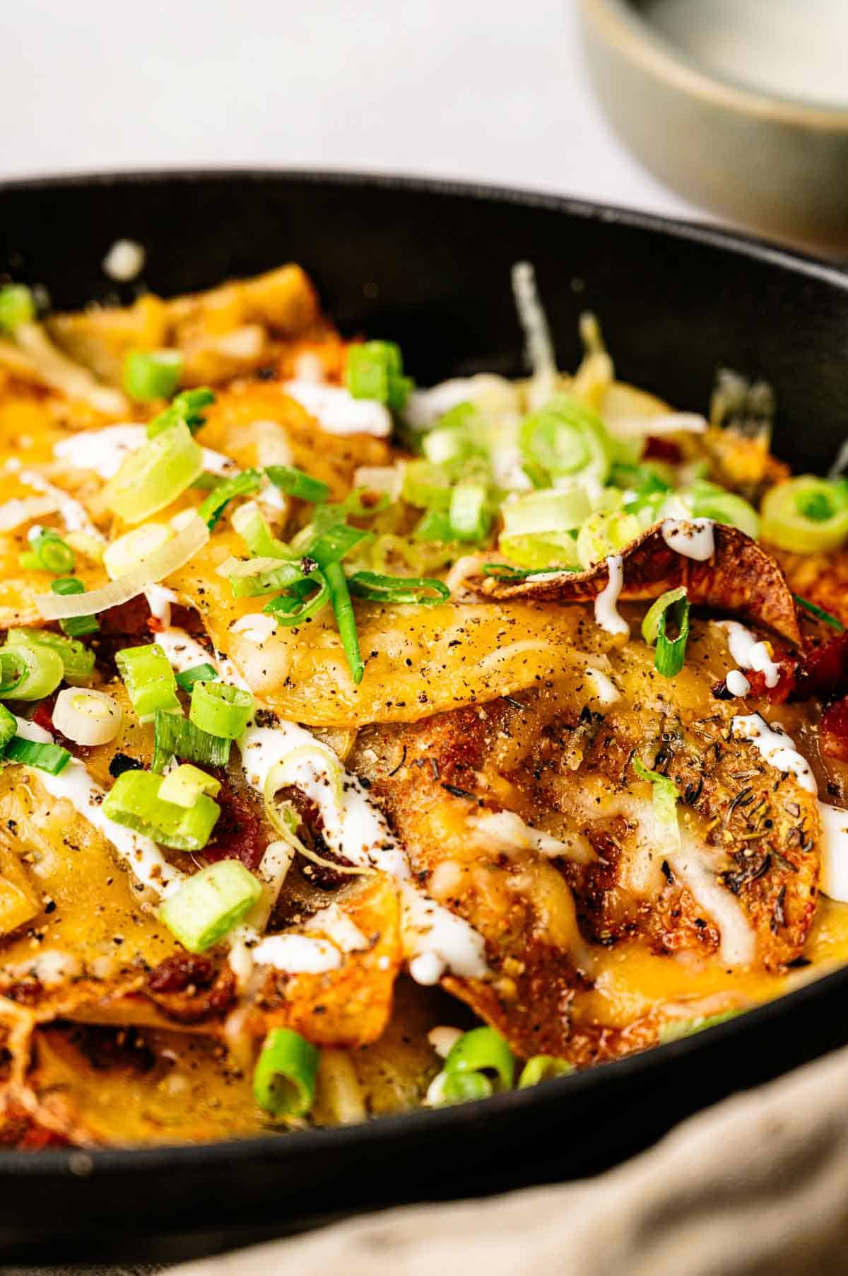 A close-up of a skillet filled with Irish Nachos topped with melted cheese, sliced green onions, black pepper, and a drizzle of white sauce.