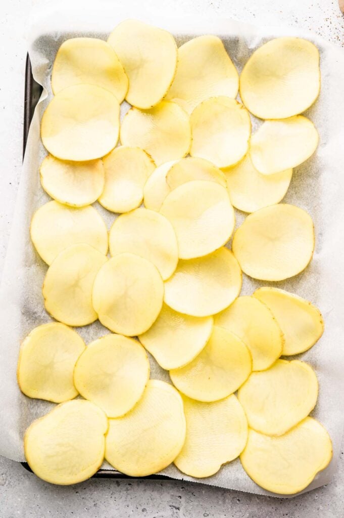 A baking tray lined with parchment paper holds several thinly sliced raw potato rounds arranged in a single layer, ready to be transformed into delicious Irish Nachos.