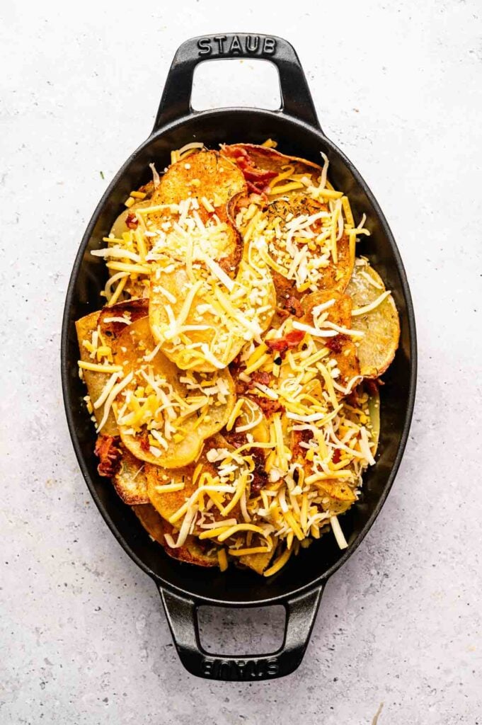 Black oval baking dish filled with sliced potatoes, crispy bacon pieces, and shredded cheese on top, ready to be baked—these Irish Nachos are the perfect comfort food twist on a classic snack.