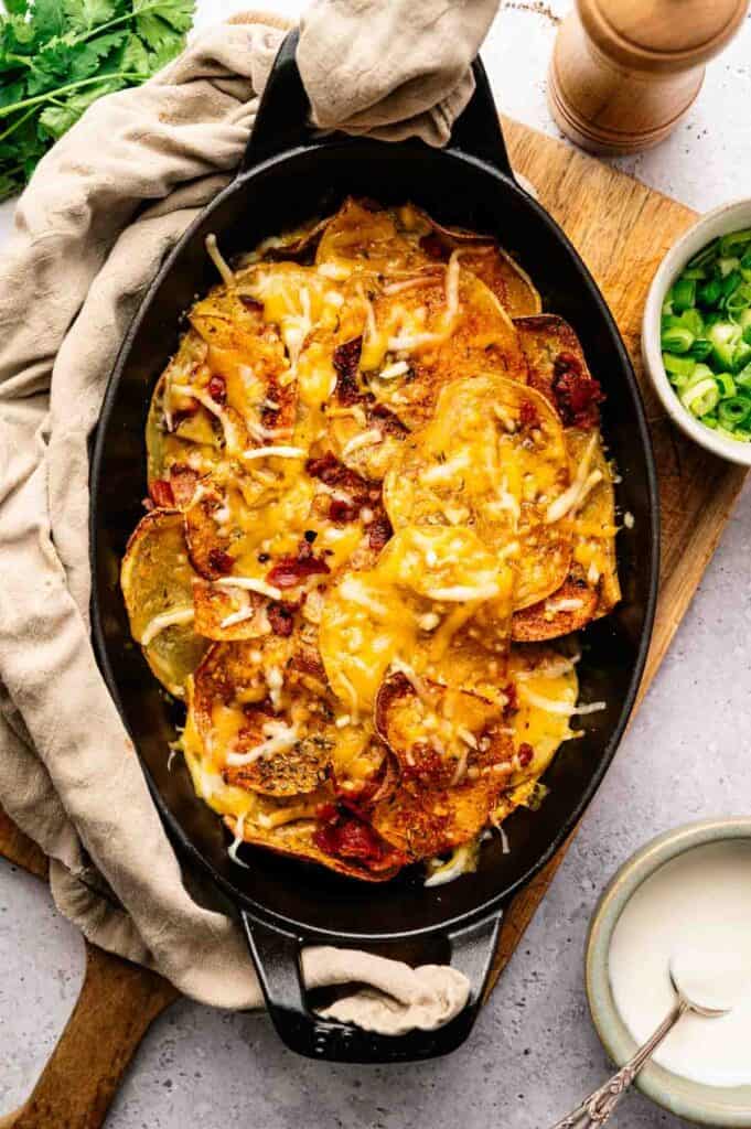 An oval baking dish filled with cheesy baked potato slices, topped with bacon bits, sits next to a bowl of chopped green onions and a small bowl of white sauce&mdash;classic Irish Nachos style.