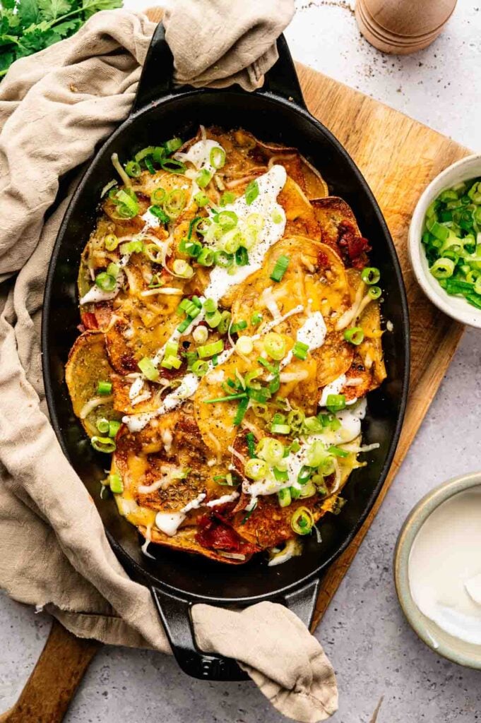 A black dish filled with layered cheesy potatoes, sliced green onions, and a drizzle of white sauce—these mouthwatering Irish Nachos are placed on a wooden board with a beige cloth and small bowls nearby.