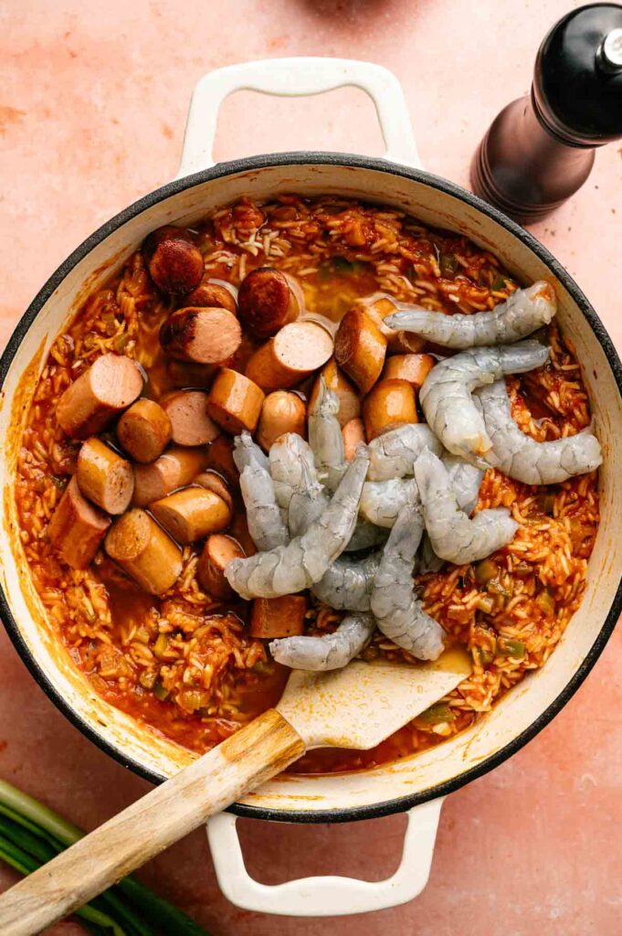 A pot of jambalaya with sliced sausage and raw shrimp nestled on top of rice in a rich, tomato-based sauce, with a wooden spoon resting inside.