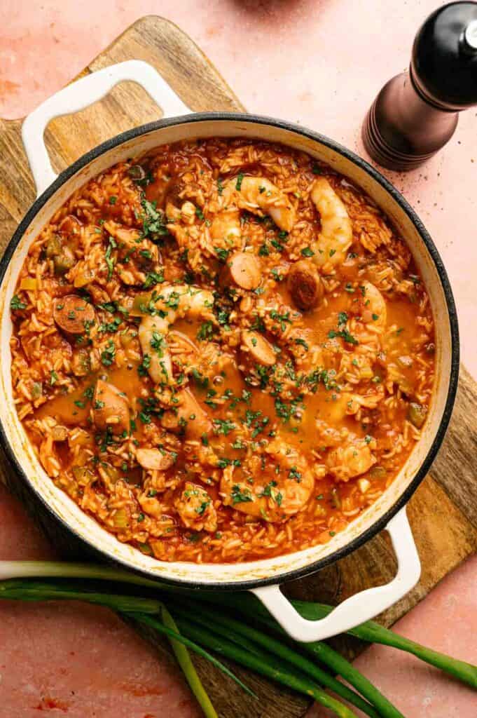 A pot of jambalaya, a seafood and rice stew, is garnished with chopped herbs and sits on a wooden board with green onions and a pepper grinder nearby.