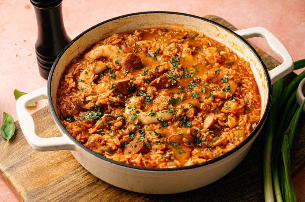 A pot of flavorful jambalaya with rice, shrimp, sausage, chicken, and vegetables, garnished with chopped herbs, sits on a wooden board.