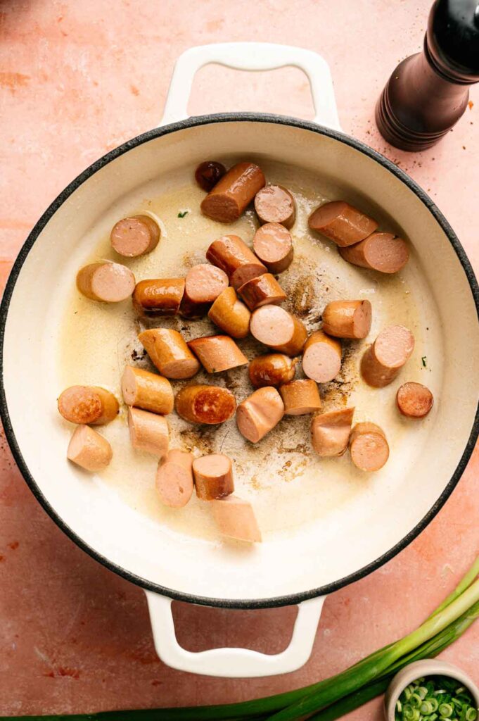 Sliced sausages are being browned in a white pot on a pink countertop, with a pepper grinder and green onions nearby—perfect beginnings for a flavorful jambalaya.