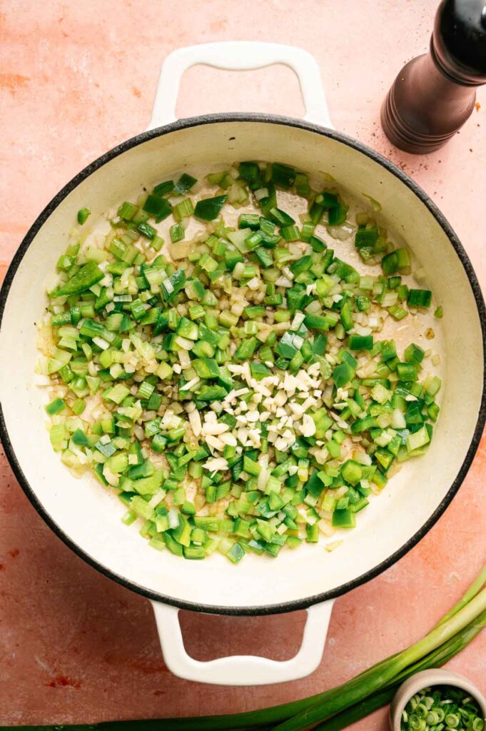 Chopped green bell peppers, onions, and minced garlic sauté in a white pot on a pink surface as the savory base for jambalaya, with a pepper grinder and a small bowl of green onions nearby.