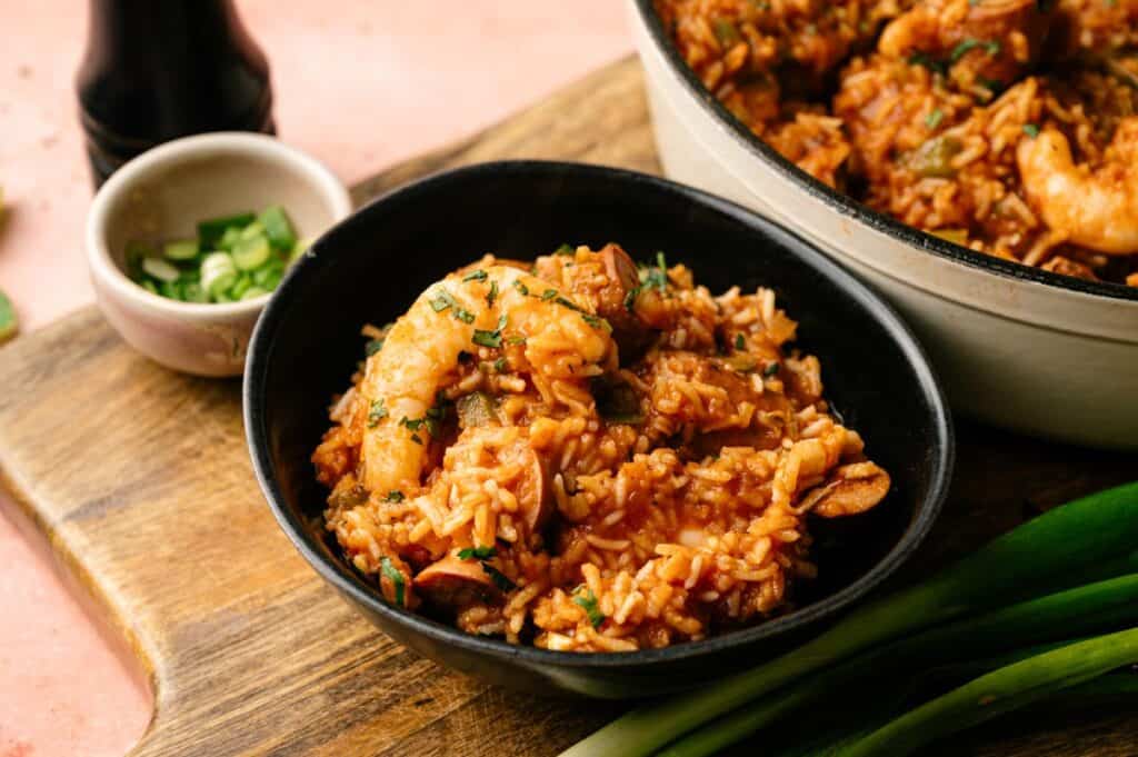 A black bowl filled with classic jambalaya&mdash;rice, shrimp, sausage, and vegetables&mdash;sits on a wooden board next to a small dish of sliced green onions.