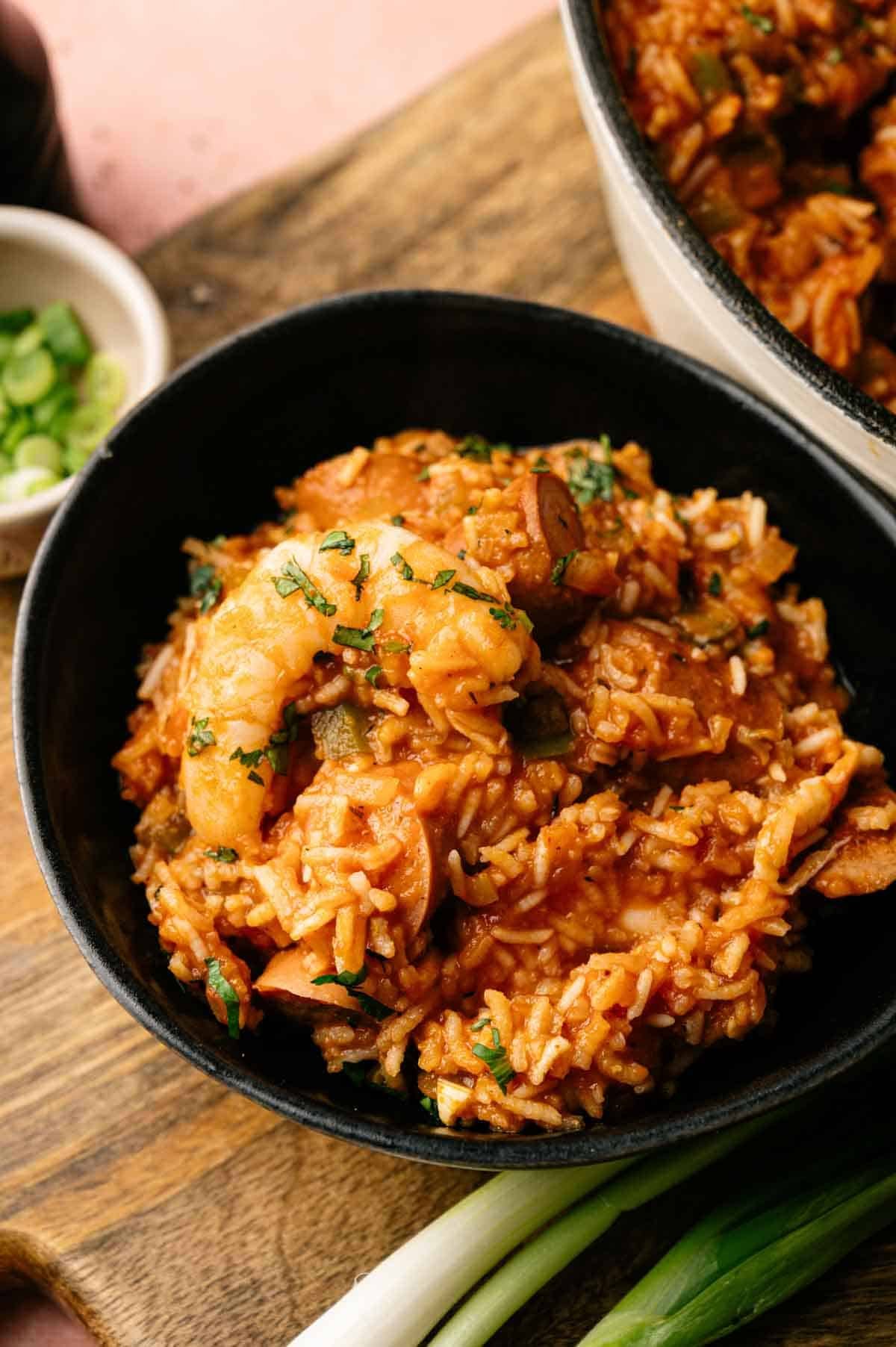 A bowl of spicy jambalaya with shrimp, sausage, rice, and vegetables is served on a wooden board and garnished with chopped herbs.