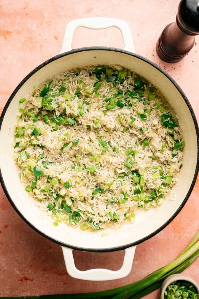 A white pot filled with cooked rice mixed with chopped green bell peppers, reminiscent of a simple jambalaya, sits on a pink surface next to a pepper grinder and a small bowl.