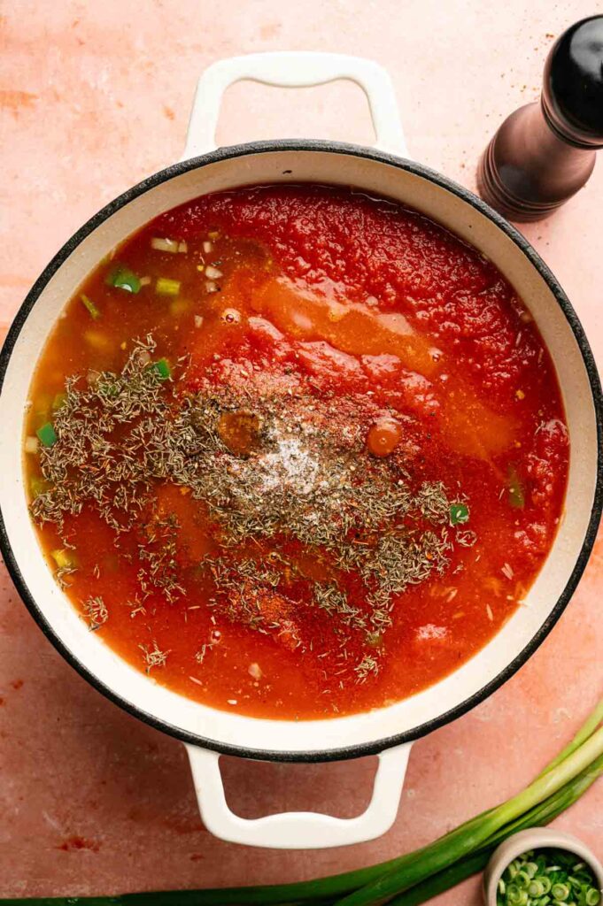 Large pot filled with tomato sauce, chopped green onions, and dried herbs—ideal for starting a flavorful jambalaya—sitting on a light pink countertop next to a pepper grinder and a small bowl of green onions.