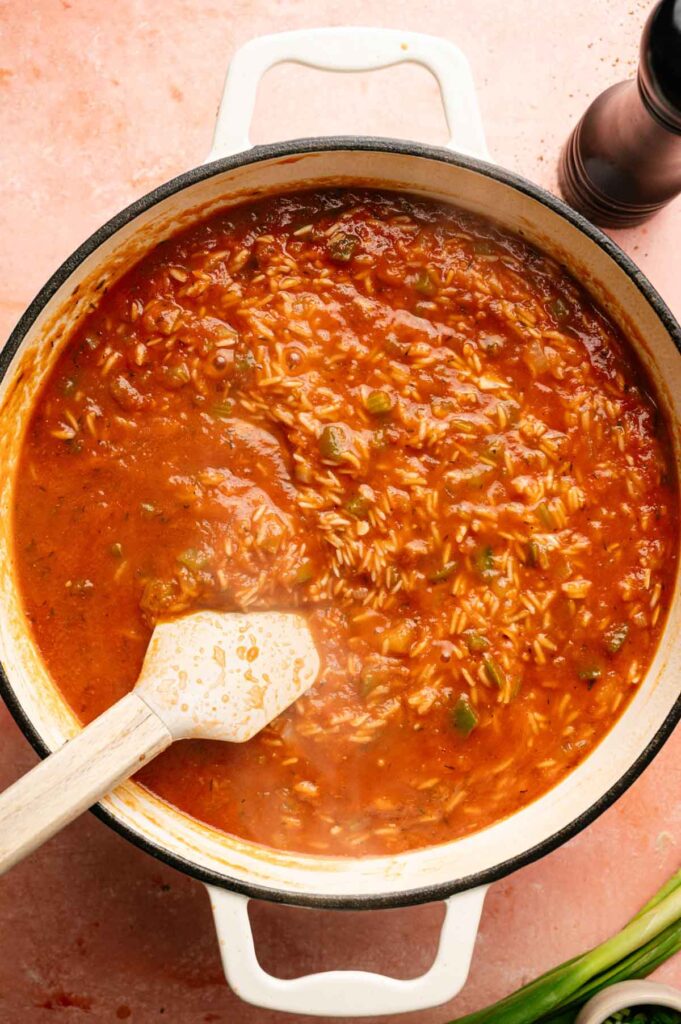 A pot of tomato-based Jambalaya rice being stirred with a wooden spoon, on a pink surface next to green onions and a black pepper grinder.