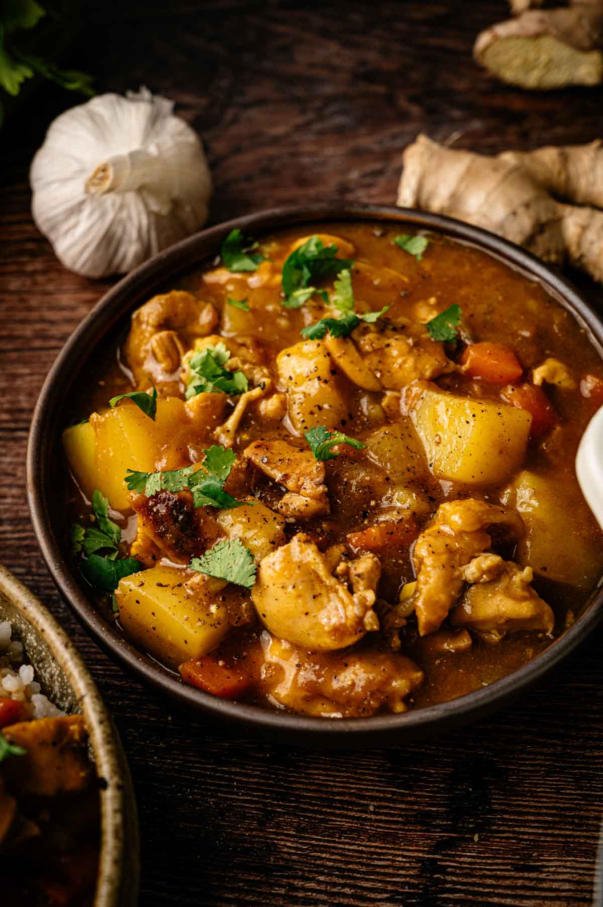 A bowl of Japanese Chicken Curry with potatoes, carrots, and fresh cilantro, placed on a wooden surface with garlic and ginger in the background.