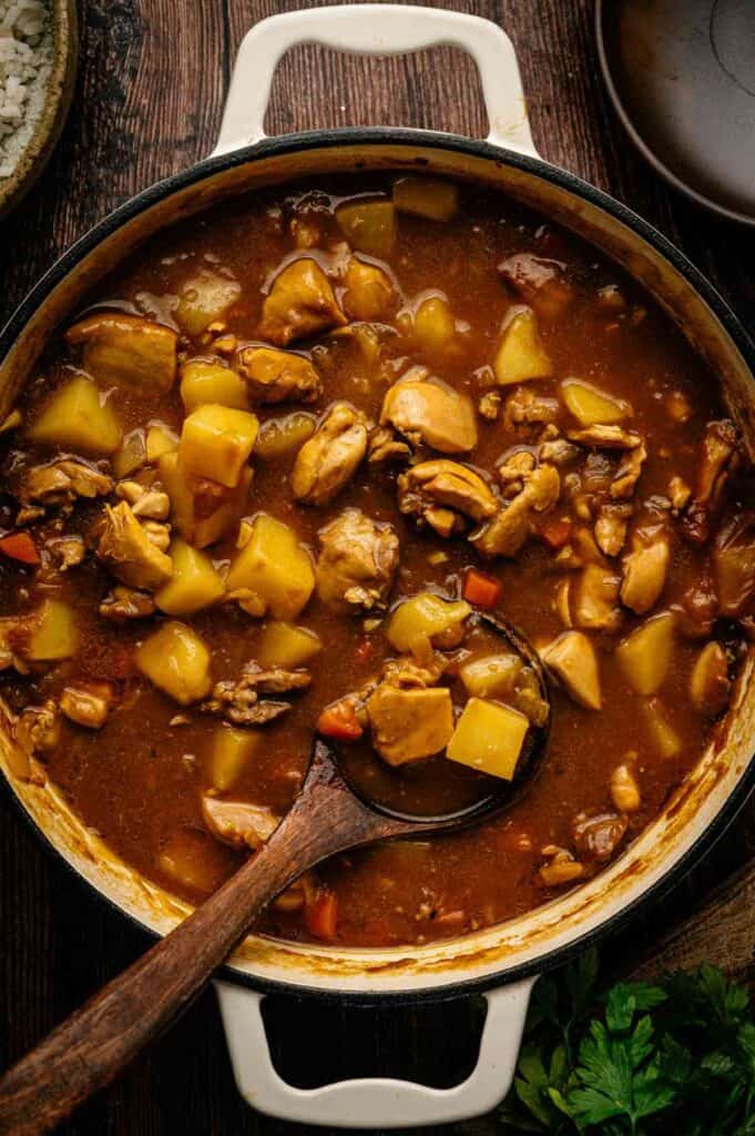 A pot of Japanese Chicken Curry with chunks of meat, potatoes, and vegetables in a thick brown broth, with a wooden ladle resting inside.