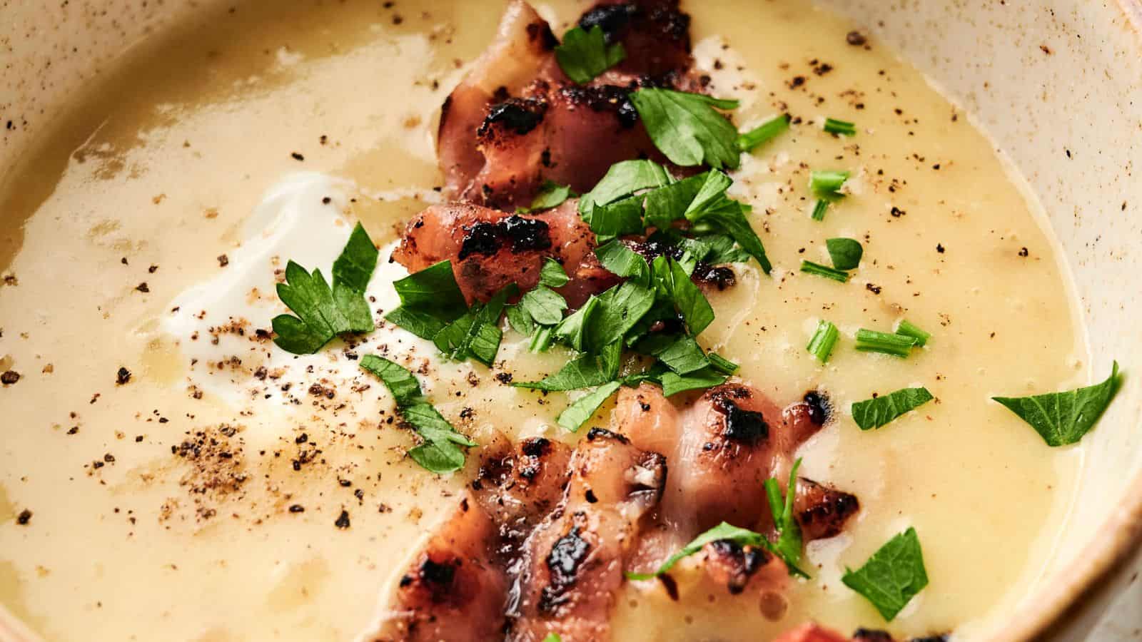 A bowl of creamy soup topped with grilled bacon pieces, chopped parsley, chives, and cracked black pepper.