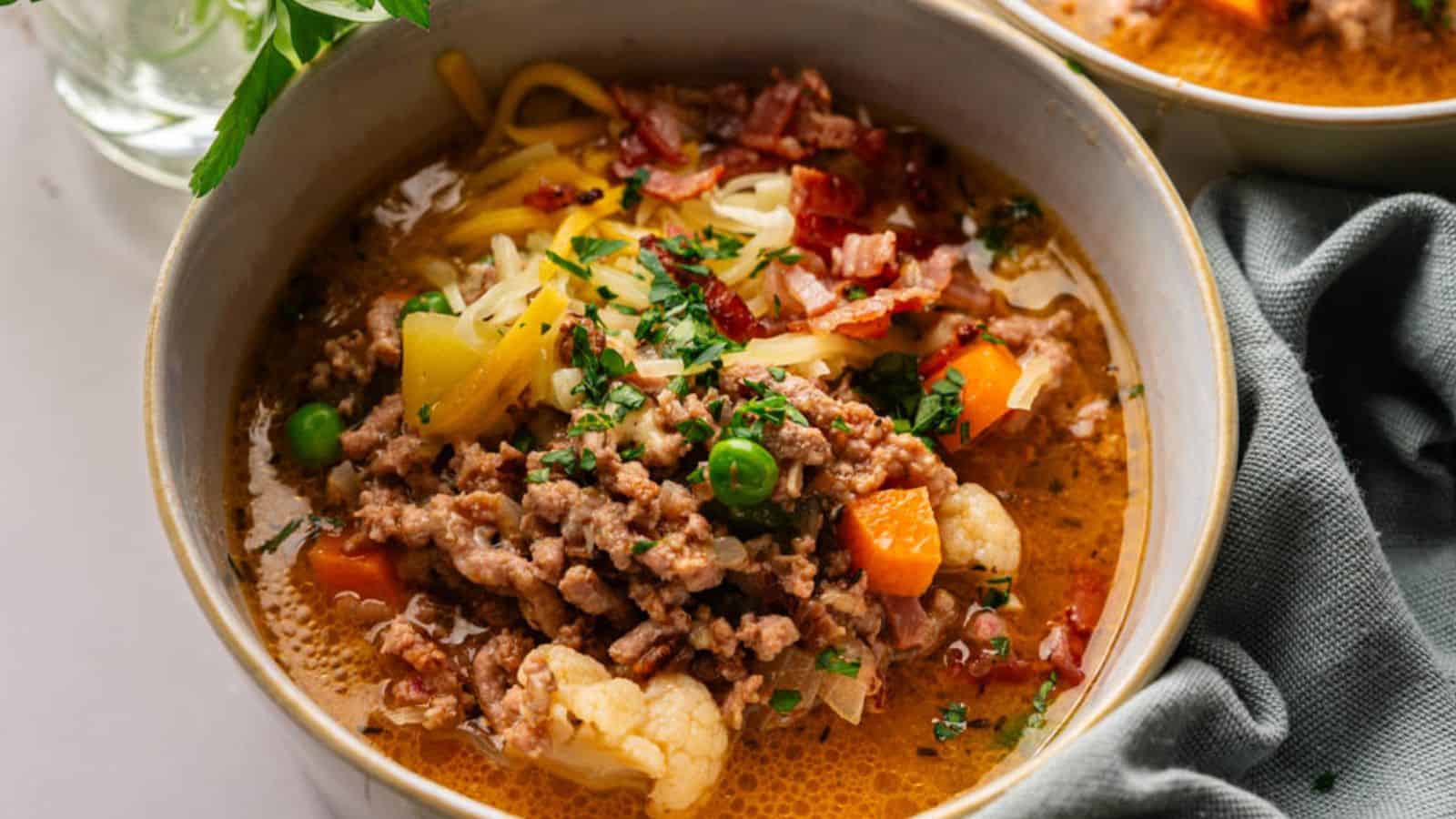 A bowl of soup with ground meat, peas, carrots, cauliflower, shredded cheese, chopped herbs, and bacon pieces, served with a napkin on the side.